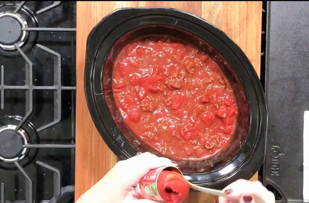 A person spooning tomato paste from a can into a crockpot filled with a red tomato-based sauce. The crockpot is placed on a wooden surface next to a stove with black burners.