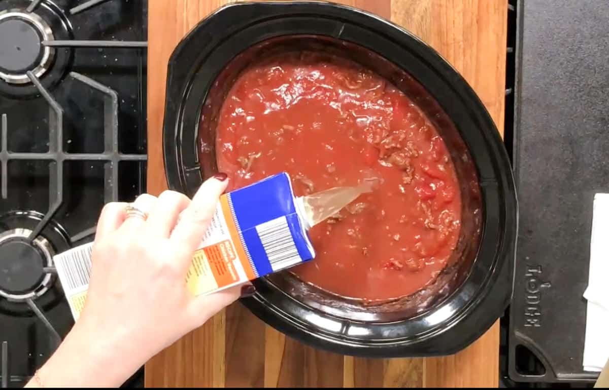 A hand is pouring broth from a carton into a black slow cooker filled with tomato-based sauce or stew. The slow cooker is on a wooden countertop next to a stove.