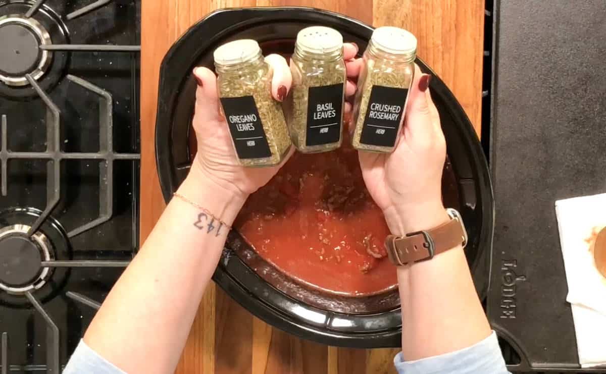 A person holds three spice containers labeled "Oregano Leaves," "Basil Leaves," and "Crushed Rosemary" over a slow cooker on a stovetop. The slow cooker contains a red sauce. A tattoo with the numbers "1213" is visible on the person's wrist.