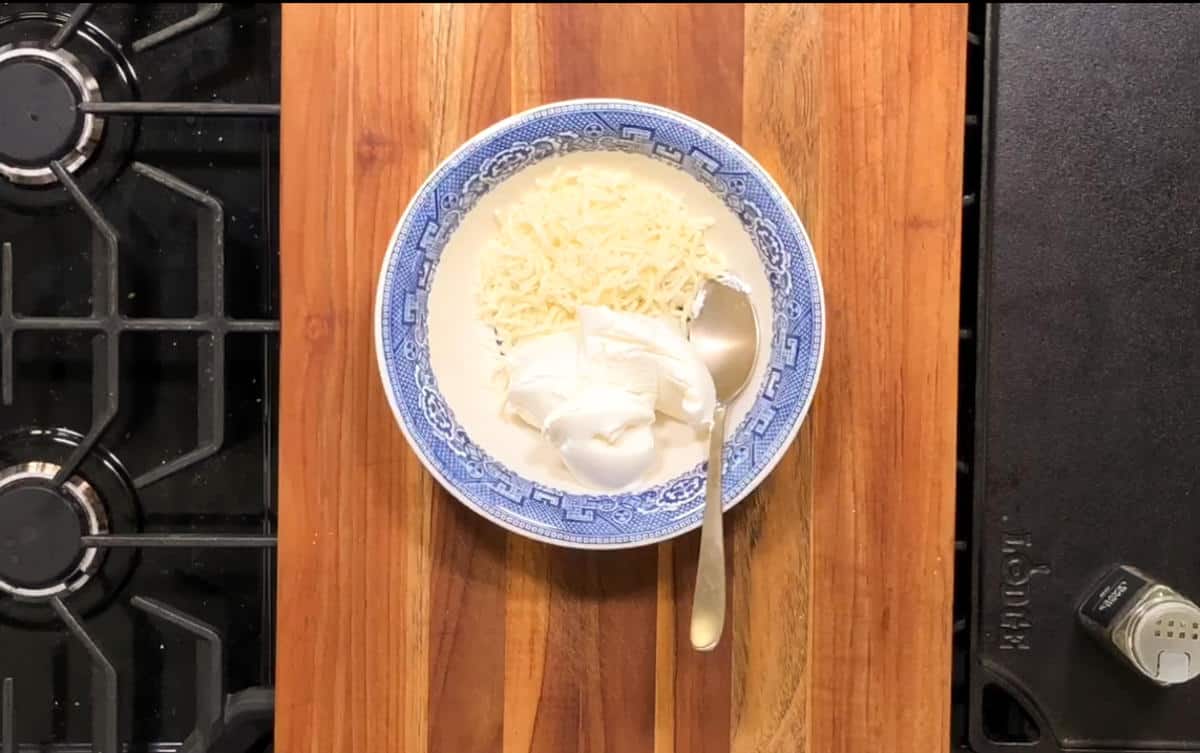 A blue and white patterned bowl containing shredded cheese and a dollop of sour cream is placed on a wooden countertop. A silver spoon rests inside the bowl. A portion of a stovetop is visible on the left and right sides of the image.