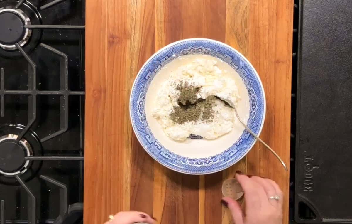 A bowl with a blue-and-white pattern containing a mixture of ingredients, including a creamy substance and sprinkles of pepper, is placed on a wooden surface near a stovetop. A person with visible hands is adding more pepper from a pepper shaker.