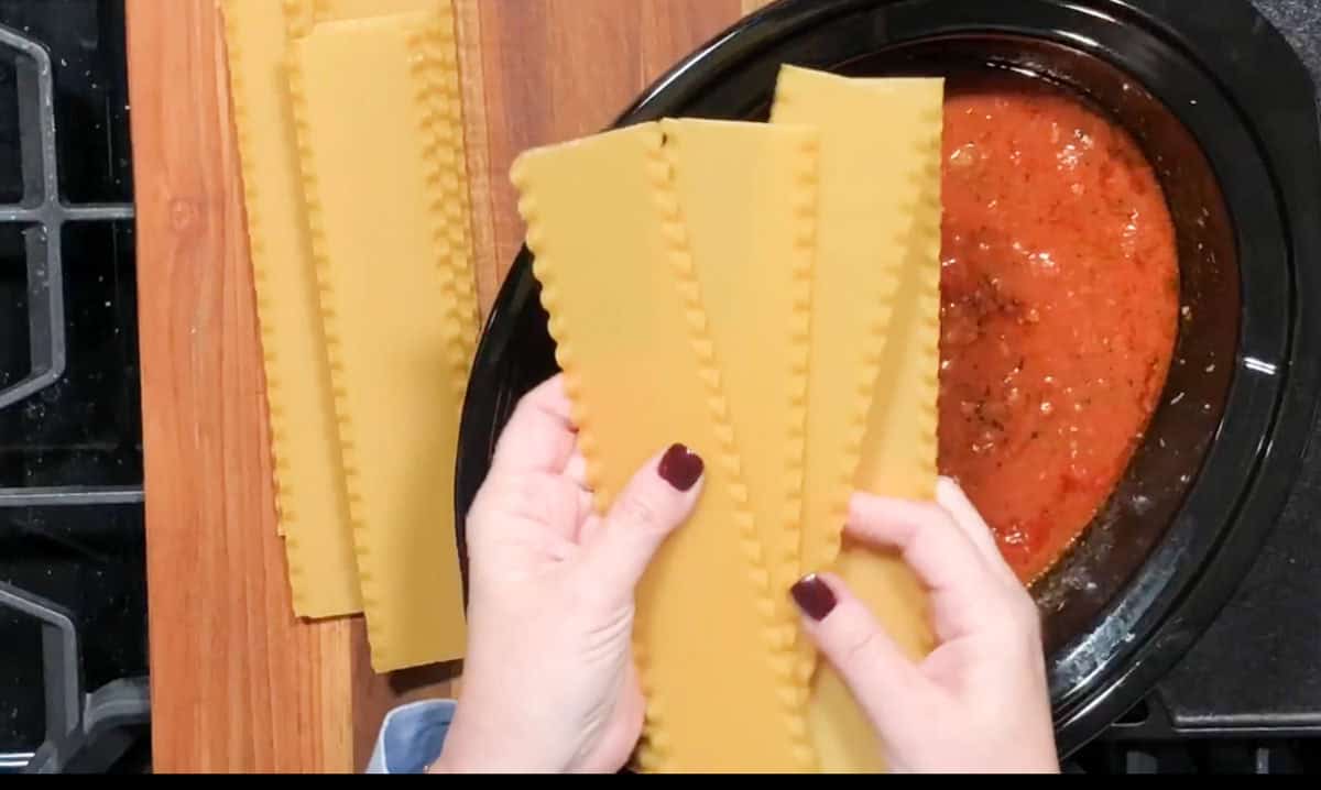 A person holding uncooked lasagna sheets over an oval slow cooker filled with tomato sauce. Several more lasagna sheets are placed on a wooden surface to the left. The person's nails are painted a dark color. A stove is partially visible in the left background.