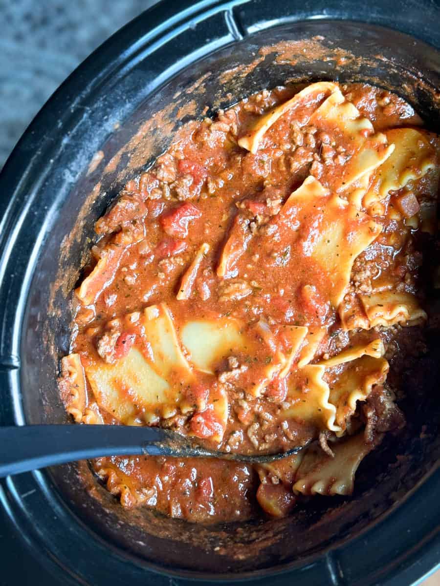 A slow cooker filled with a mixture of lasagna noodles, ground meat, tomato sauce, and spices. A serving spoon is placed inside the pot, and the contents appear to be thoroughly mixed and cooked.