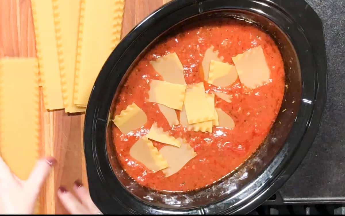 A black slow cooker is filled with red pasta sauce and layered with broken pieces of lasagna noodles. On the left, there are uncooked lasagna noodles resting on a wooden countertop, with a person's hands visible near them.