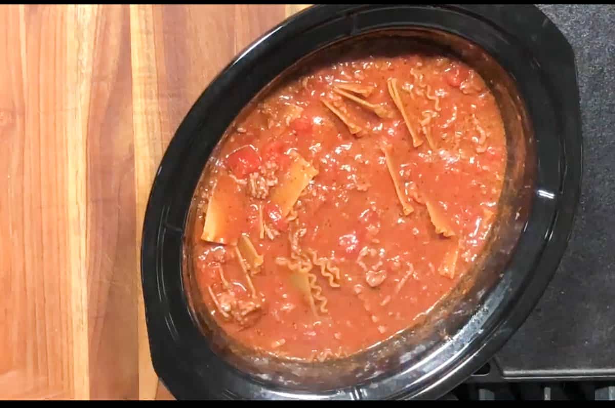 A black slow cooker on a wooden countertop is filled with a red tomato-based sauce containing broken pieces of lasagna noodles, ground meat, diced tomatoes, and seasonings. The dish appears to be a lasagna soup or stew.