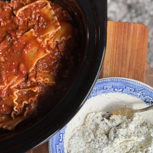 A slow cooker filled with lasagna is on the left side of the image. On the right, there's a blue and white plate with a portion of ricotta cheese and a spoon resting in it. The scene is set on a wooden surface.