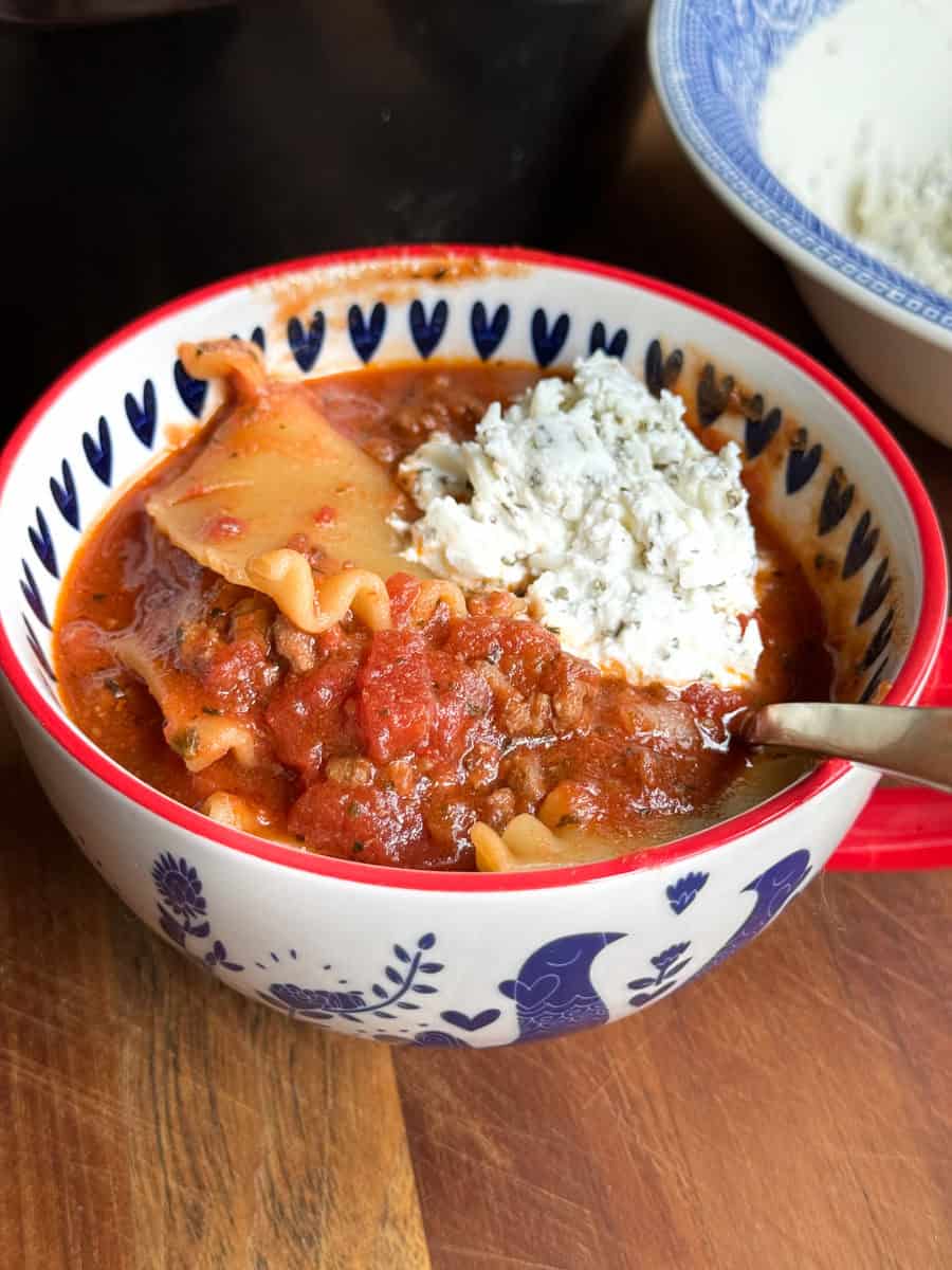 A bowl of lasagna soup is shown with a red and white patterned bowl. The soup includes lasagna noodles, a rich tomato sauce with meat, and a generous dollop of ricotta cheese topped with herbs. A spoon rests inside the bowl.