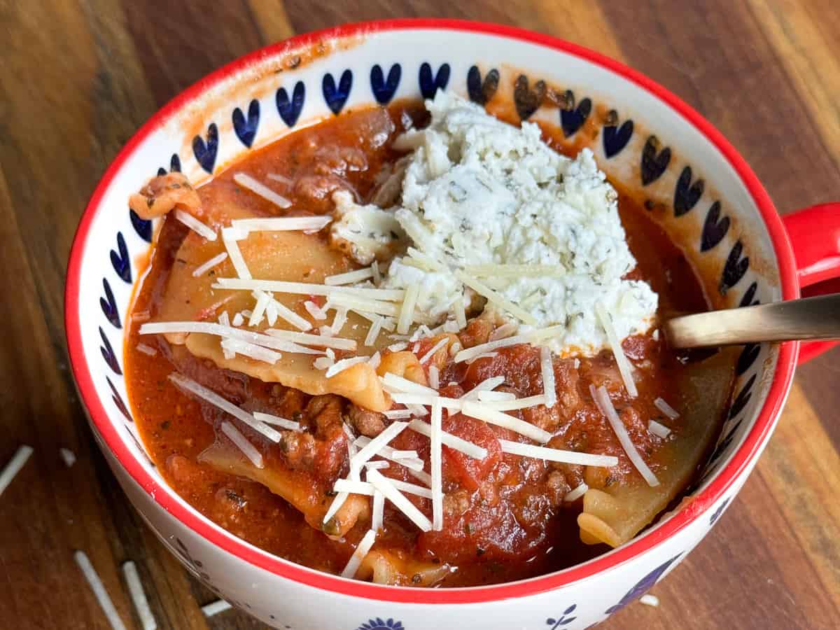 A bowl filled with lasagna soup, with pieces of lasagna noodles, ground meat, and tomato sauce. The soup is topped with shredded Parmesan cheese and a dollop of ricotta cheese. The bowl has a red rim and patterned sides, and a spoon is resting inside.