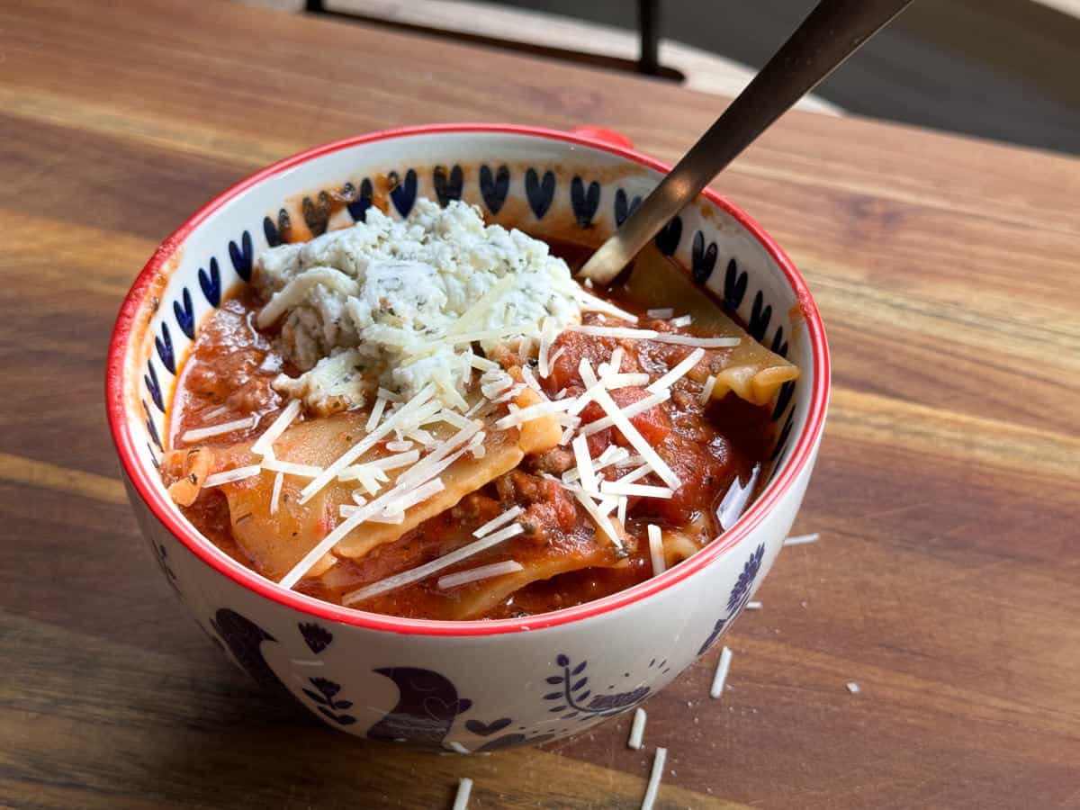 A bowl of lasagna soup sits on a wooden surface. The soup is topped with shredded cheese and appears to contain chunks of lasagna noodles and a tomato-based sauce. A spoon is placed inside the bowl, and the bowl has a decorative pattern.