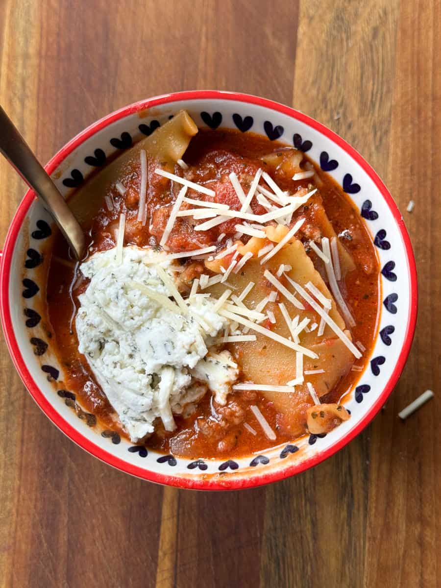 A bowl containing lasagna soup topped with a dollop of ricotta cheese and shredded parmesan. The soup features pieces of lasagna noodles in a rich tomato-based sauce. The bowl is white with a red rim and black heart pattern, with a spoon inside.