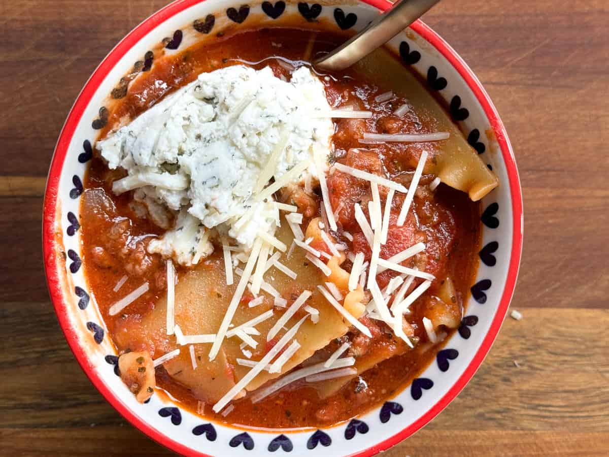 A bowl of lasagna soup with large pasta pieces, tomato-based broth, and a dollop of ricotta cheese topped with shredded parmesan. The bowl has a red rim and heart-shaped patterns and is placed on a wooden surface. A spoon rests inside the bowl.