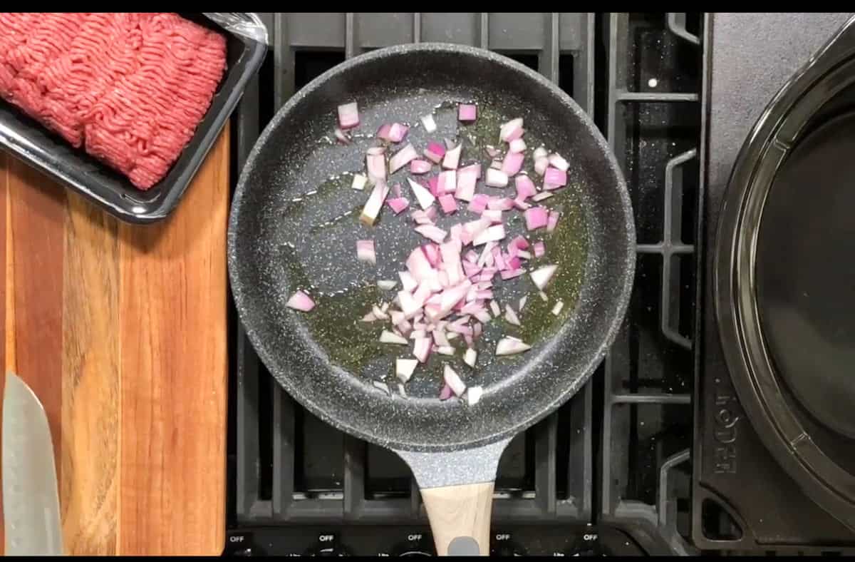 A saucepan containing chopped red onions is being heated on a stove. Beside the pan, there is a package of raw ground meat and a large knife on a wooden cutting board. The stove has several cooking settings visible.