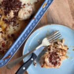 A serving of a cheesy baked dish with ground meat is placed on a blue plate beside a fork and knife. The baking dish, also containing the dish, rests on a wooden surface.