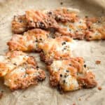 Cheese crisps sprinkled with black and white sesame seeds are scattered on a piece of parchment paper. The crisps have a golden-brown color and a textured, crispy appearance.