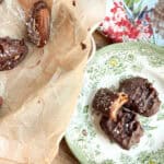 A plate with chocolate-covered dates sprinkled with sea salt on a floral-patterned dish. Nearby, a parchment paper holds more dates. A floral cloth is partially visible in the corner.