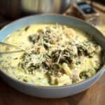 A bowl of creamy soup is filled with ingredients such as ground meat, leafy greens, sliced potatoes, and a sprinkle of grated cheese on top. A spoon rests in the soup, and the background includes a blurry spice container and a pot.
