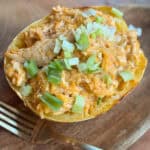 A halved spaghetti squash filled with a mixture of cooked rice, shredded chicken, and topped with chopped green onions is placed on a wooden surface next to a gold fork.