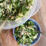 A bowl of kale salad with shredded cheese and nuts is placed on a wooden surface next to a larger container of the same salad. There are serving utensils in both the bowl and the container.