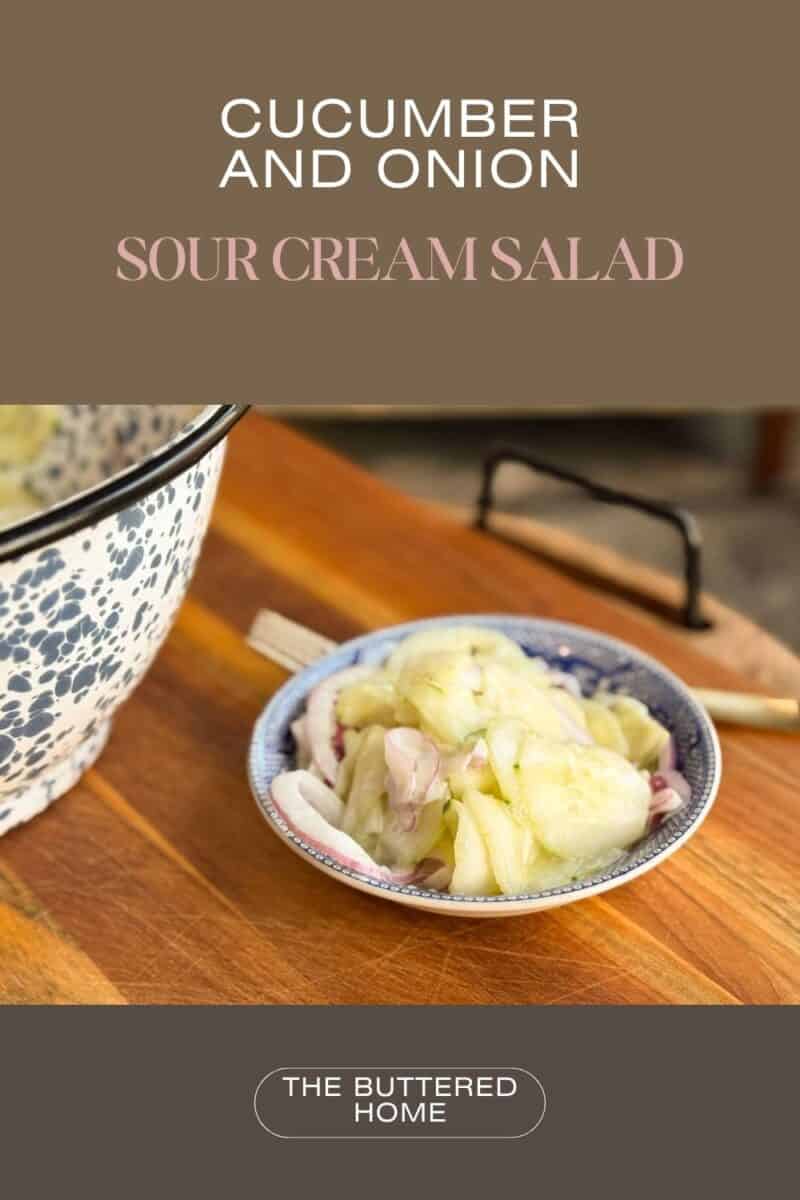 Close-up of a blue and white bowl containing cucumber and onion salad on a wooden table. The text reads, "Cucumber and Onion Sour Cream Salad" and "The Buttered Home" with a larger bowl partially visible on the left.