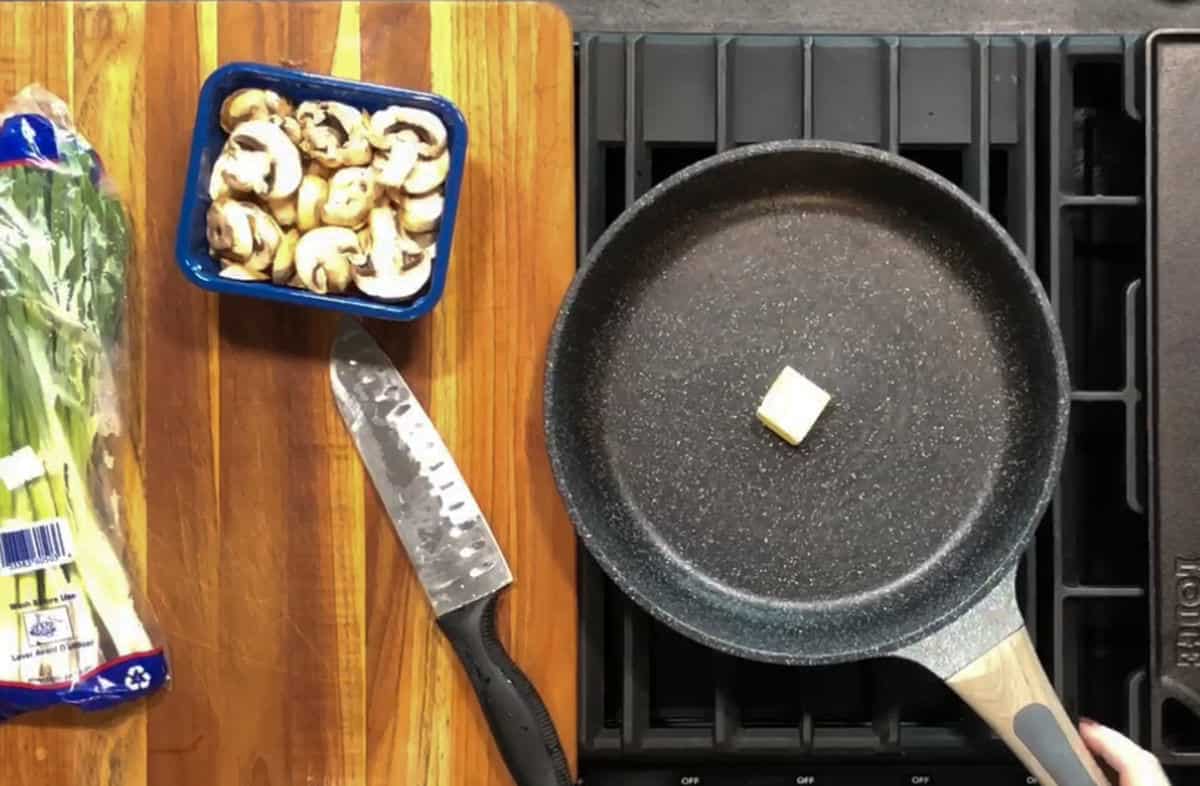A frying pan with a small piece of butter sits on a stovetop. Next to it, a wooden cutting board holds a knife, a container of sliced mushrooms, and a package of green onions.
