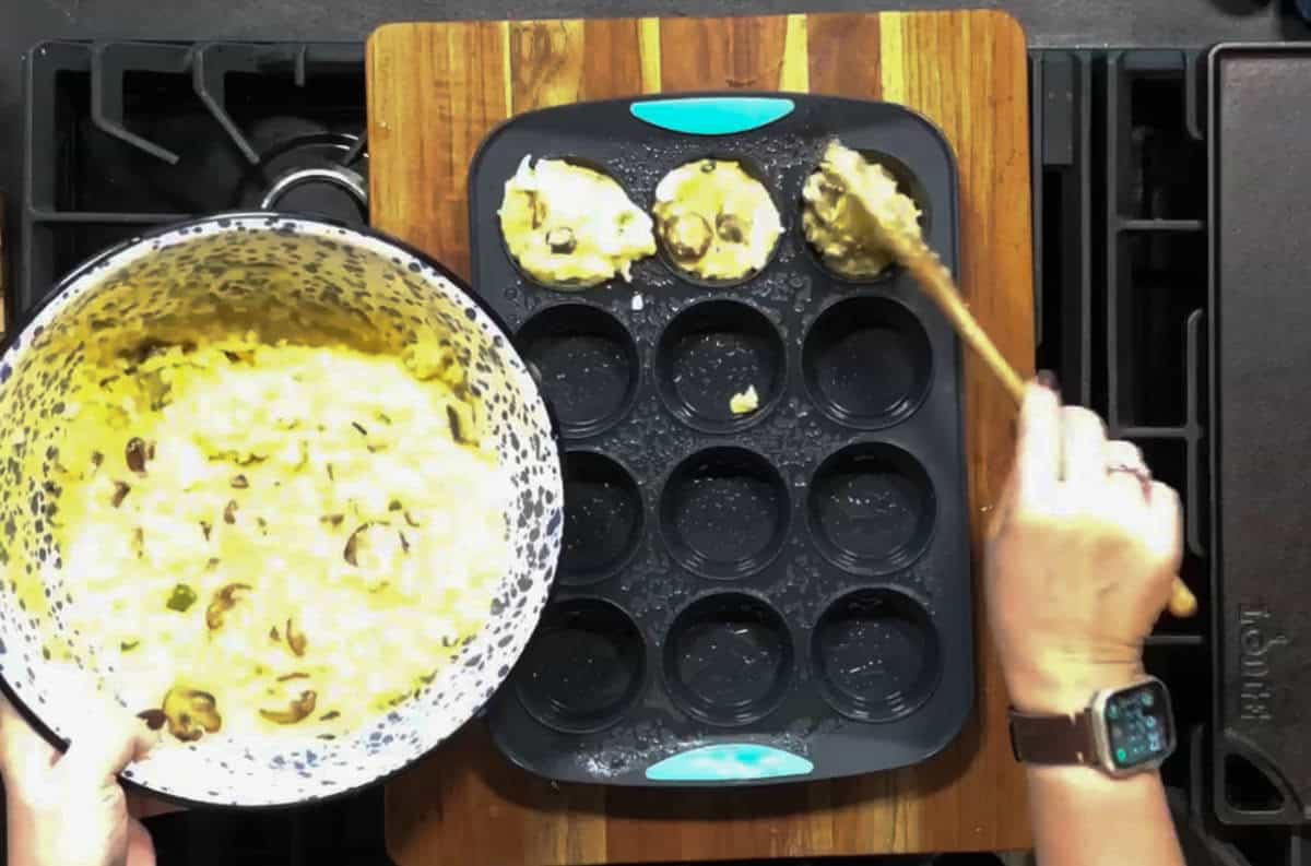 Two hands are visible, one holding a bowl with a mixture containing batter and mushrooms, while the other uses a spoon to distribute the mixture into a muffin tin with three sections filled. The muffin tin is on a wooden board on a stovetop.