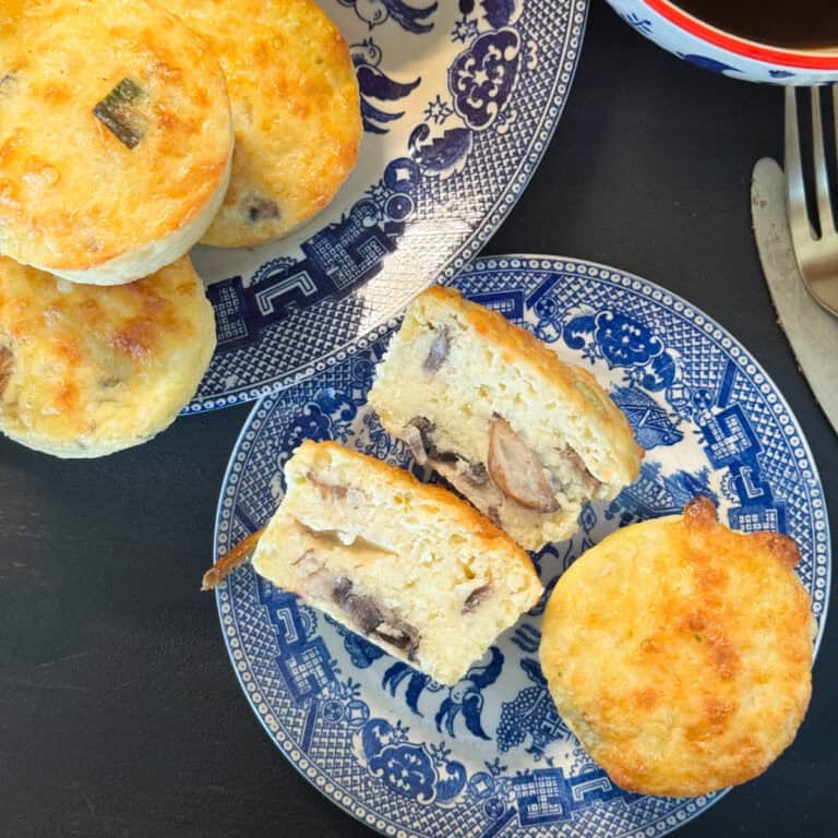 Blue and white plates with scones on a dark surface. Some scones are whole, while others are cut open, revealing a filling with visible mushroom pieces. A silver knife and fork are partially visible on the right.