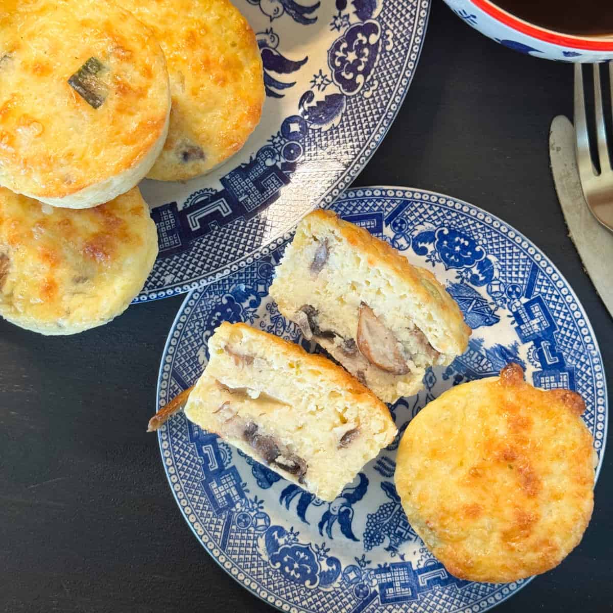 Blue and white plates with scones on a dark surface. Some scones are whole, while others are cut open, revealing a filling with visible mushroom pieces. A silver knife and fork are partially visible on the right.