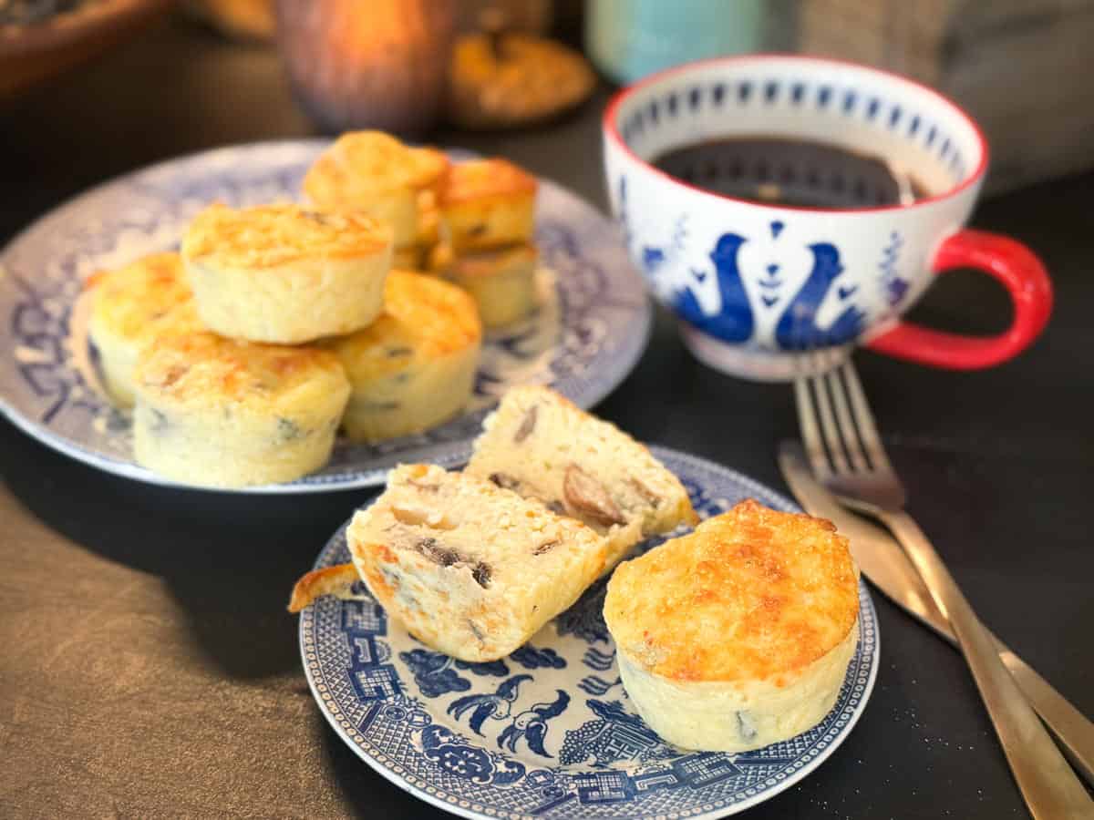 A set of small quiches and a slice are on a blue patterned plate. Another plate with more quiches is in the background. A cup of black coffee in a red-handled mug sits beside the plate. A fork and knife are placed on the table.