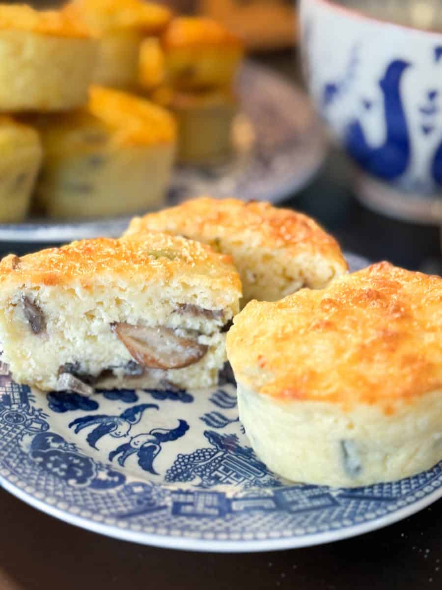 Two savory muffins are placed on a blue and white patterned plate, one is cut open showing mushroom pieces inside. More muffins and a blue and white bowl are in the background.