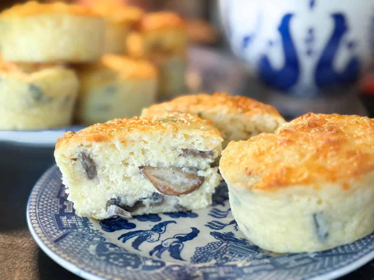 Two baked muffins with a fluffy texture and visible mushroom pieces are on a blue and white patterned plate. More muffins are blurred in the background, with a blue and white patterned mug also visible.