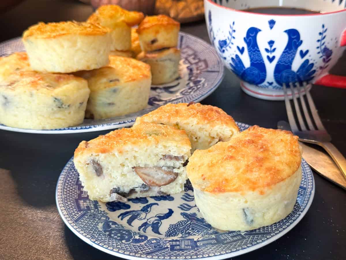 Two blue and white plates display savory muffins with visible pieces of mushroom and cheese. One plate has a partially cut muffin showing the interior. A decorative blue and white cup is in the background, alongside a red-handled fork.