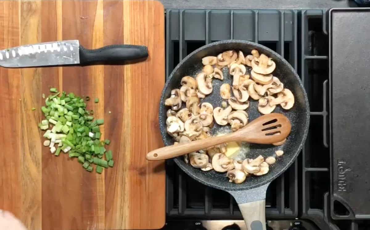 A cutting board with a knife and chopped green onions is on the left. On the right, a frying pan on a stovetop contains sautéing sliced mushrooms stirred with a wooden spoon.