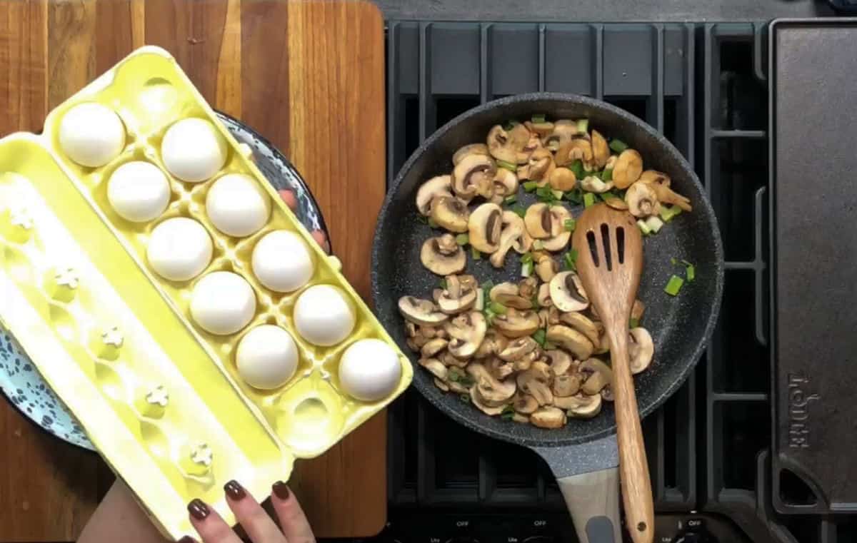 A hand holding a yellow egg carton with seven white eggs is visible. A frying pan on a stove contains sliced mushrooms and green onions. A wooden spatula rests in the pan, and the scene is set on a wooden and black stovetop surface.