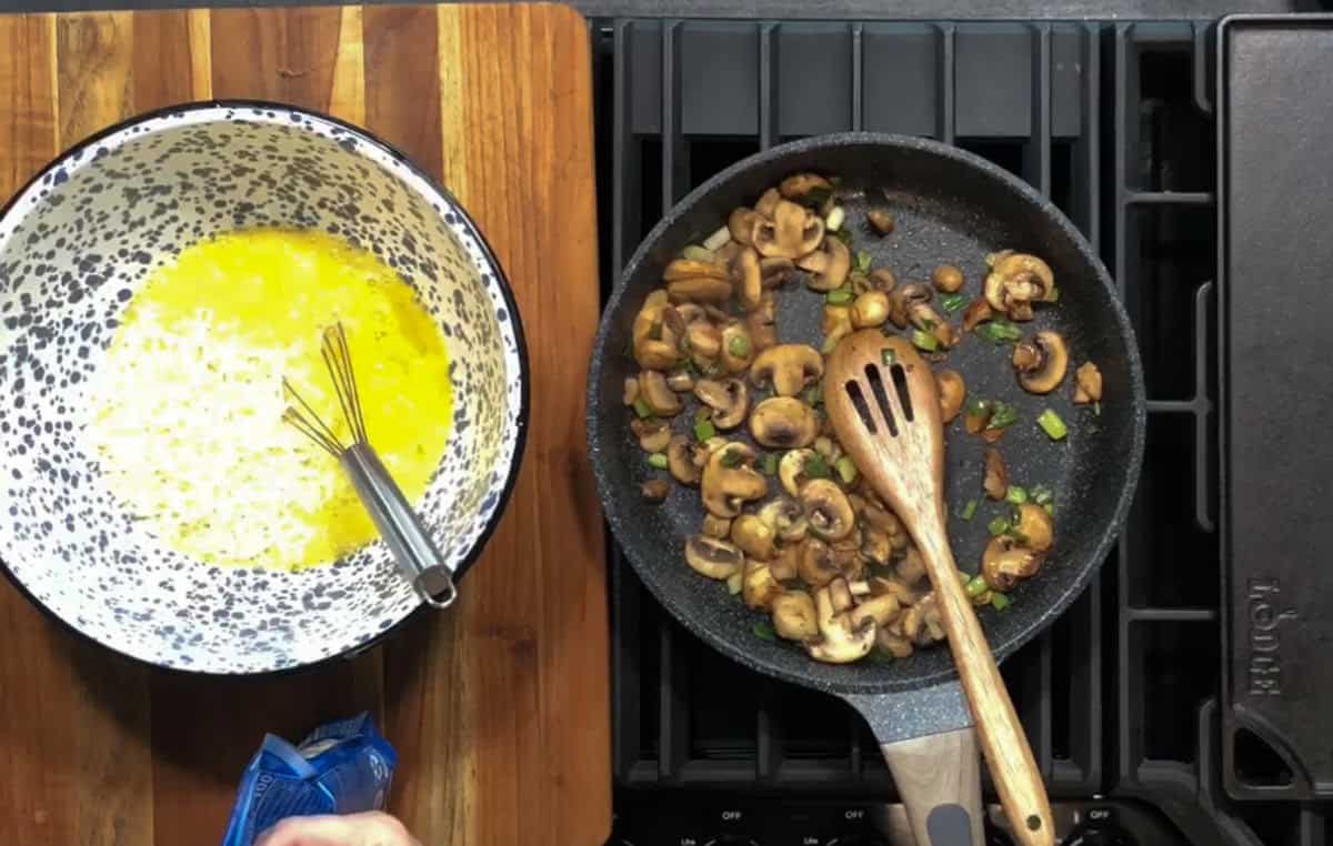 A top-down view of a kitchen counter shows a bowl with a yellow mixture and a whisk on the left, and a frying pan with cooked mushrooms and green onions on the right. A hand is holding a packet near the bottom left corner.