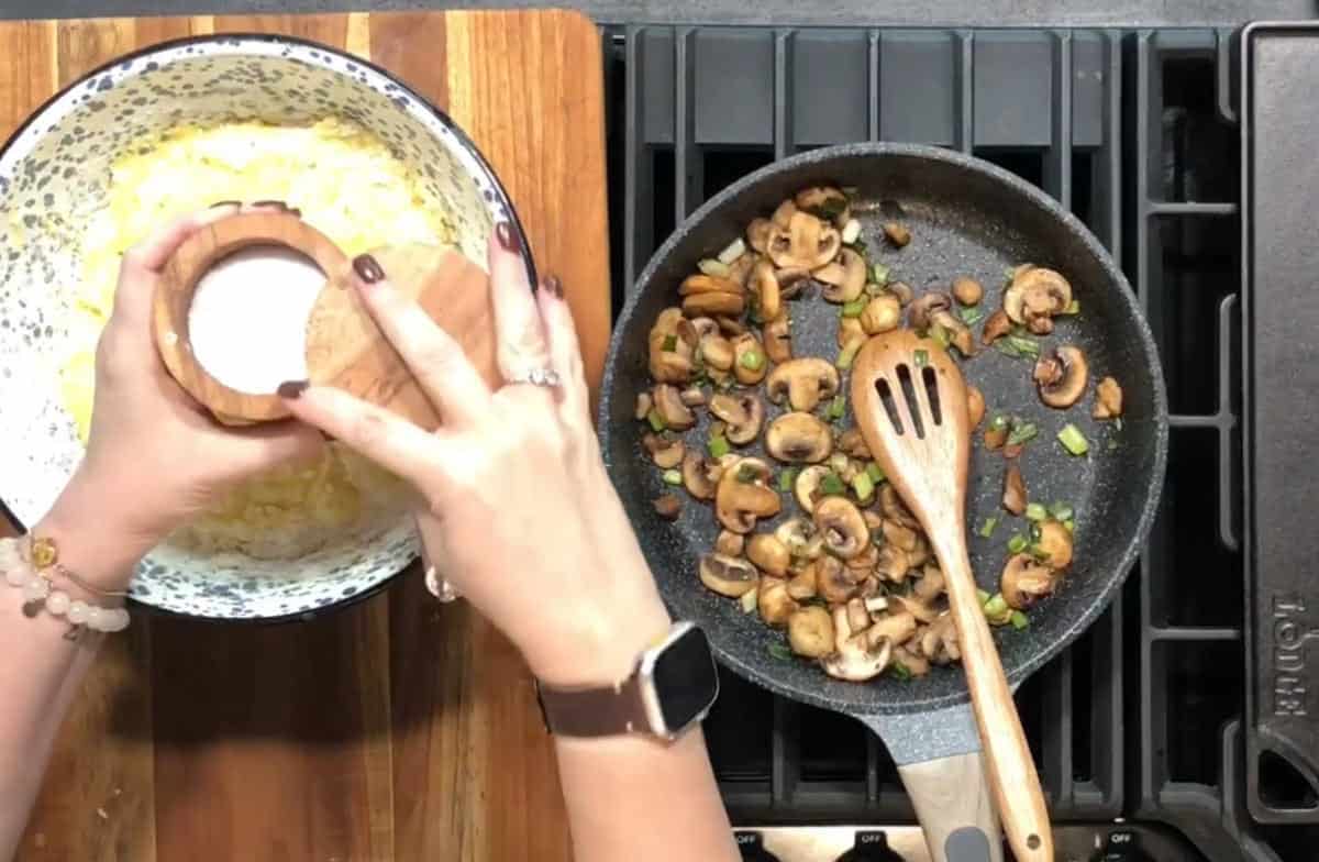 A person is adding salt to a bowl of mashed potatoes on a wooden countertop. A skillet with sautéed mushrooms and green onions is next to the bowl. A wooden spoon rests in the skillet. The person is wearing a smartwatch.