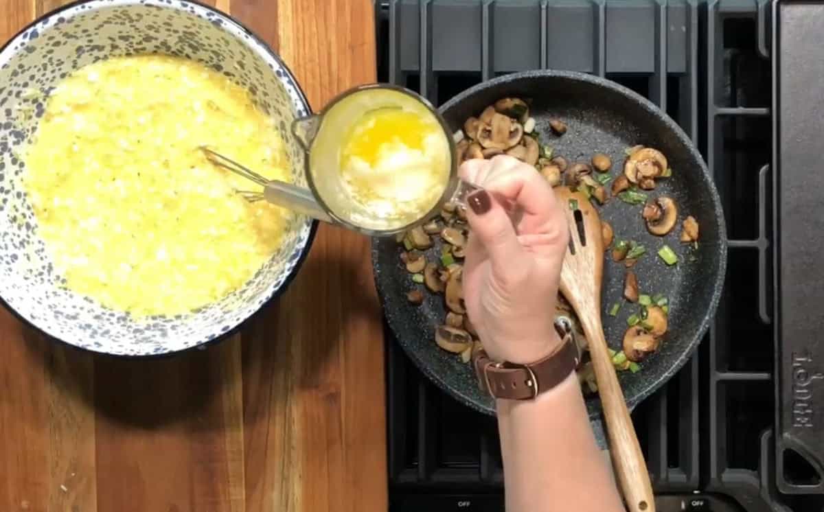 A person is cooking on a stovetop. On the left, there is a bowl with a mixture of beaten eggs. On the right, a pan contains sautéed mushrooms and green onions. The person holds a glass of melted butter above the pan.
