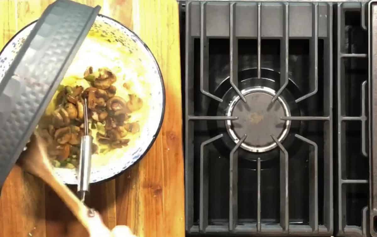 A hand holds a wooden spoon stirring a bowl of food with mushrooms on a wooden countertop next to a gas stove with an empty burner. A pan lid is partially visible on the left.