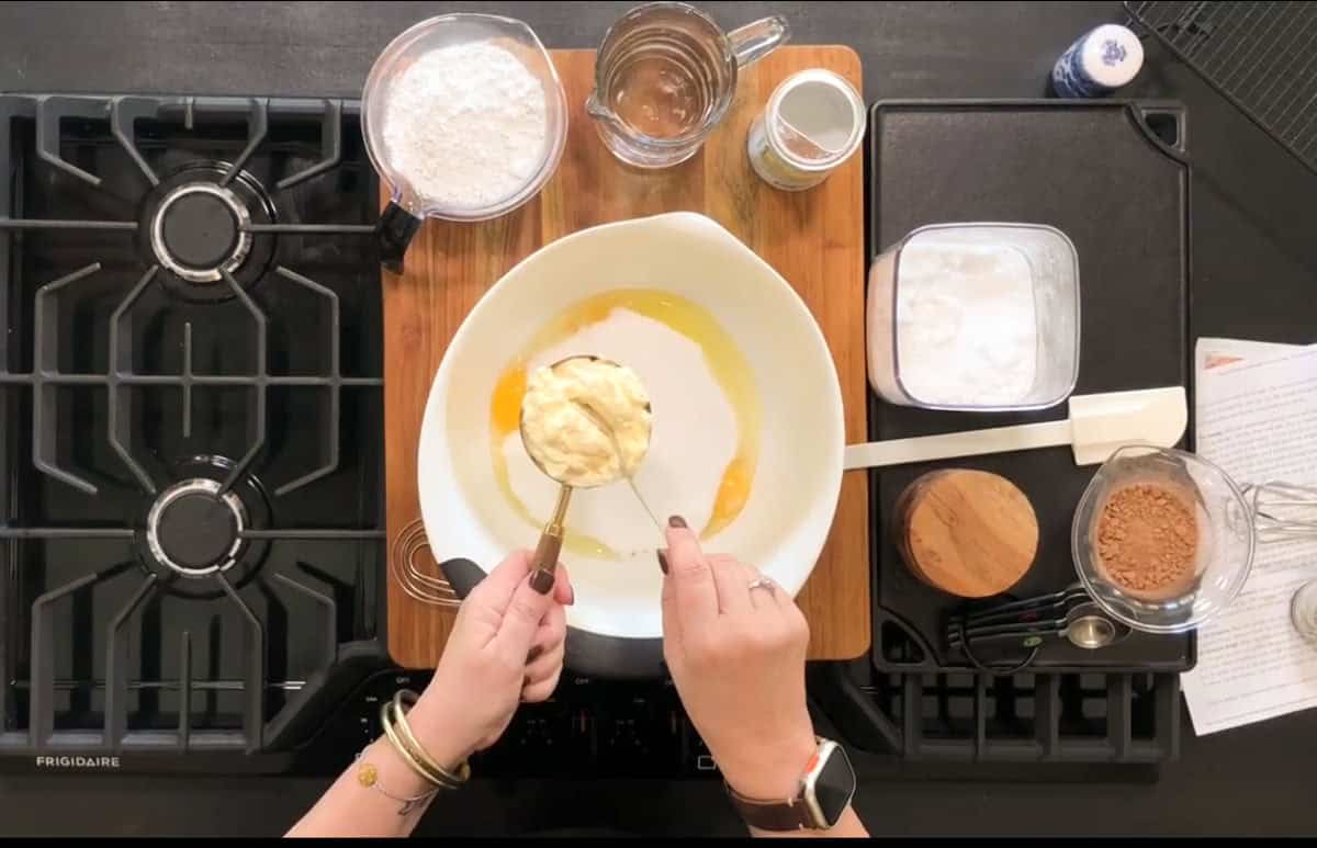A person is preparing ingredients for baking, with a white mixing bowl containing eggs and a scoop of another ingredient. Surrounding the bowl are various ingredients in small containers, such as flour and cocoa, on a wooden board over a stove.