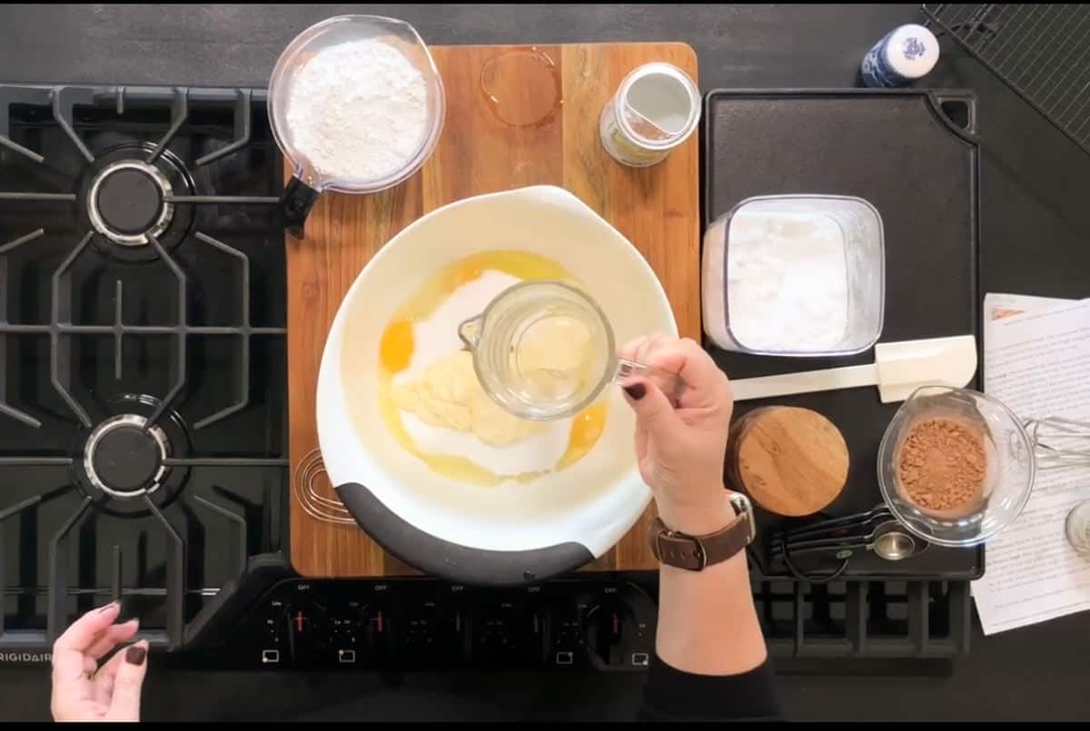 A person is pouring a liquid ingredient into a bowl containing eggs and other ingredients on a wooden board. The kitchen counter has flour, cocoa, a whisk, and a recipe sheet nearby. The setting is on a stovetop with a black surface.