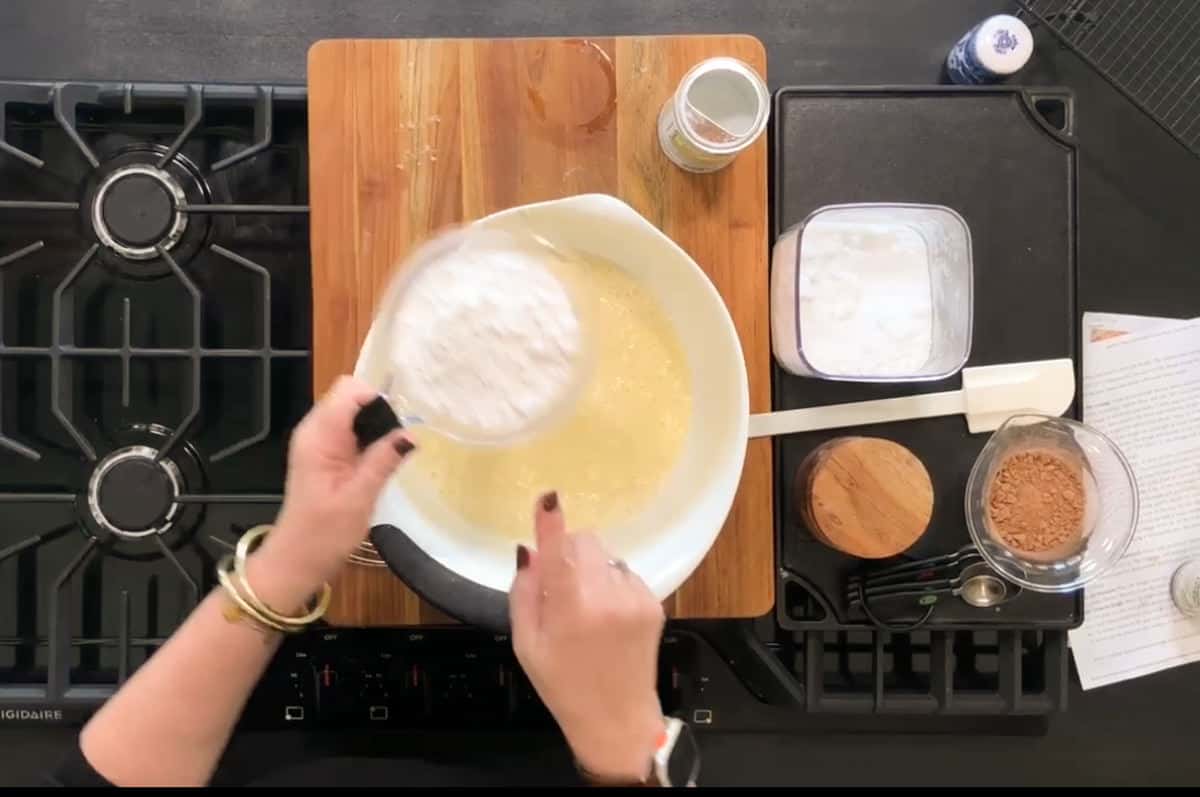 A person is holding a measuring cup filled with flour over a mixing bowl on a wooden board. The kitchen counter also has jars of ingredients, a spatula, and a sheet of paper nearby.