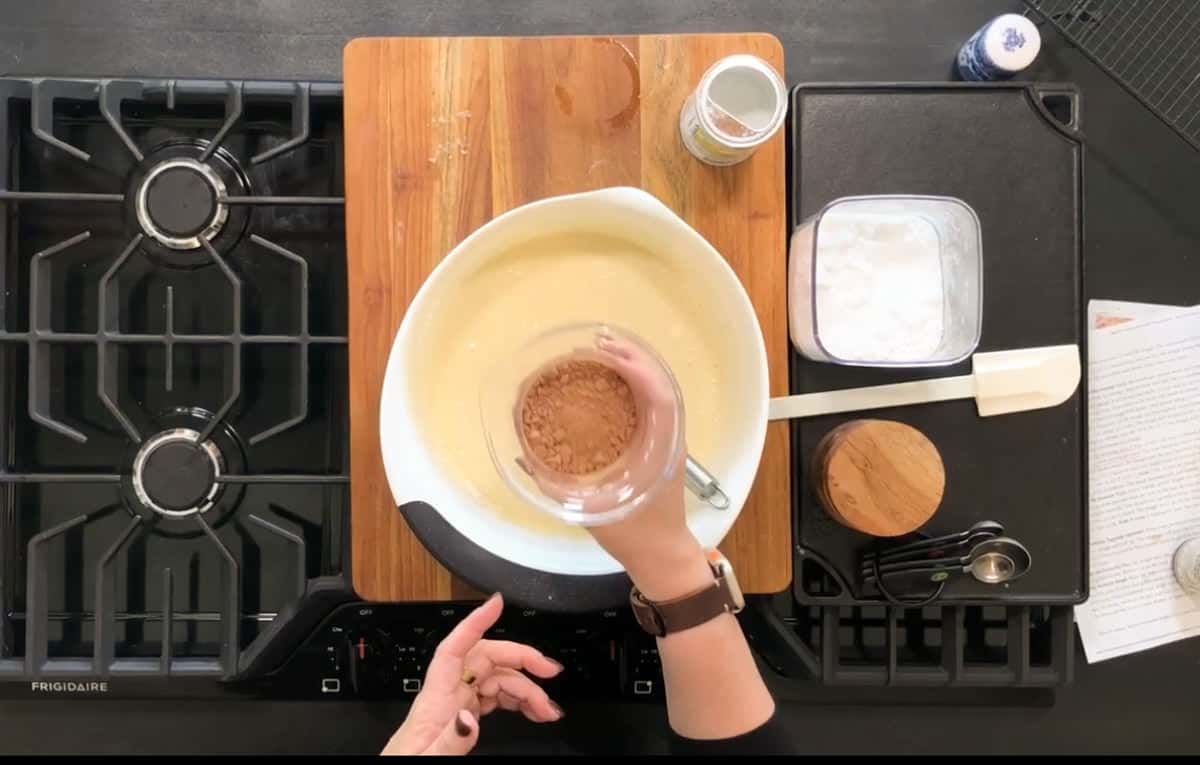 A person is holding a small glass bowl of cocoa powder over a mixing bowl containing yellow batter. The scene is set on a black stovetop with a wooden cutting board, a jar, a spatula, and measuring spoons nearby.