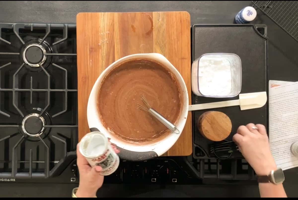 Top-down view of a kitchen counter. A person is holding a can labeled "CARNATION," pouring contents into a brown mixture in a white mixing bowl with a whisk. Nearby are a wooden cutting board, a white bowl with powder, a wooden container, and a recipe sheet.