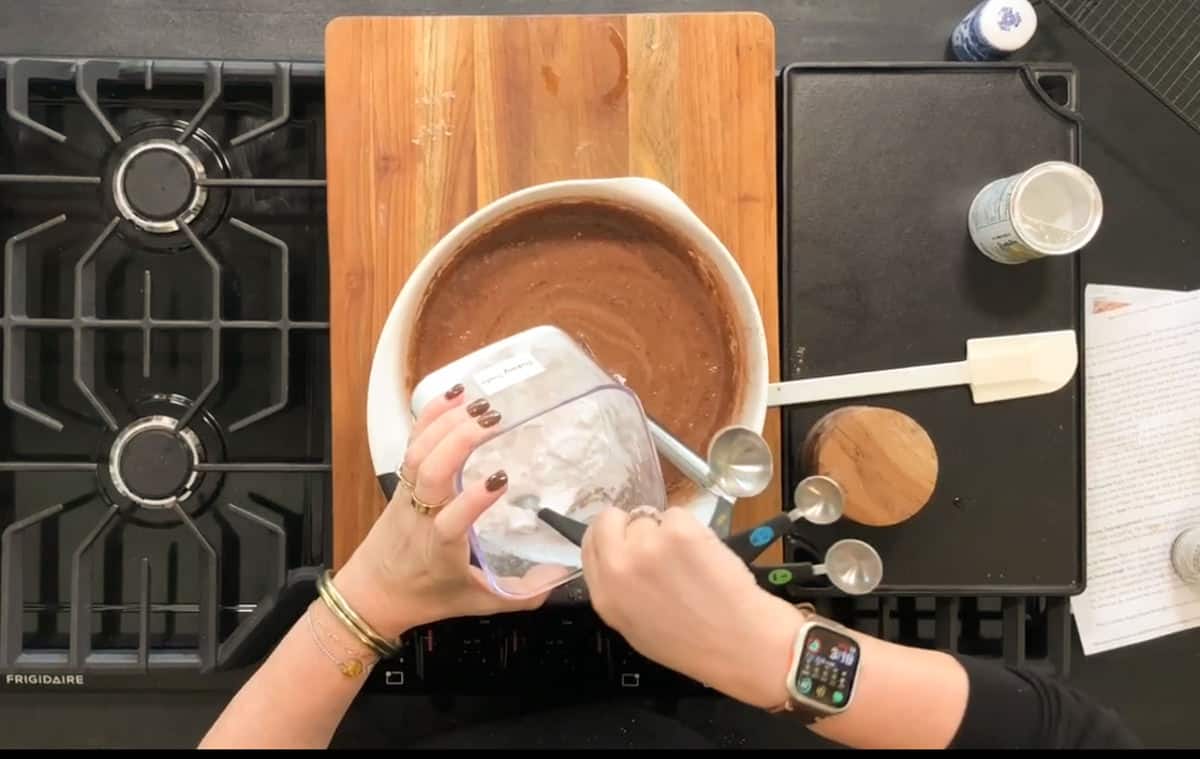 A person is preparing chocolate cake batter in a mixing bowl on a wooden cutting board. The ingredients include flour and cocoa powder. Measuring spoons and a spatula are nearby on a black stovetop. The person is wearing a smartwatch.