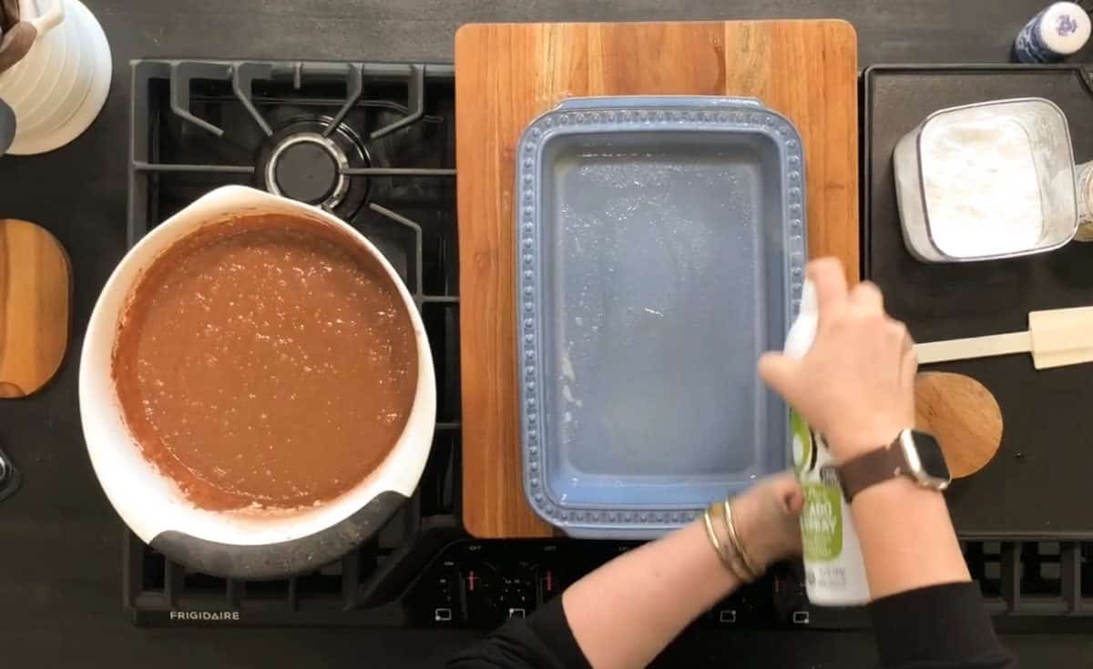 A person is spraying a blue baking dish with cooking spray. A mixing bowl filled with brown batter and a white spatula lie nearby on a black stovetop. A wooden cutting board is positioned under the baking dish. Other baking ingredients are visible.