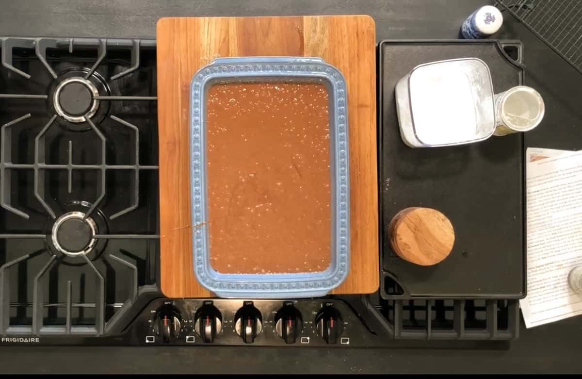 A blue baking dish filled with brown batter sits on a wooden cutting board on a black stovetop. Nearby are a glass container, a metal container with a white lid, a small wooden container, and some papers on a metal tray.