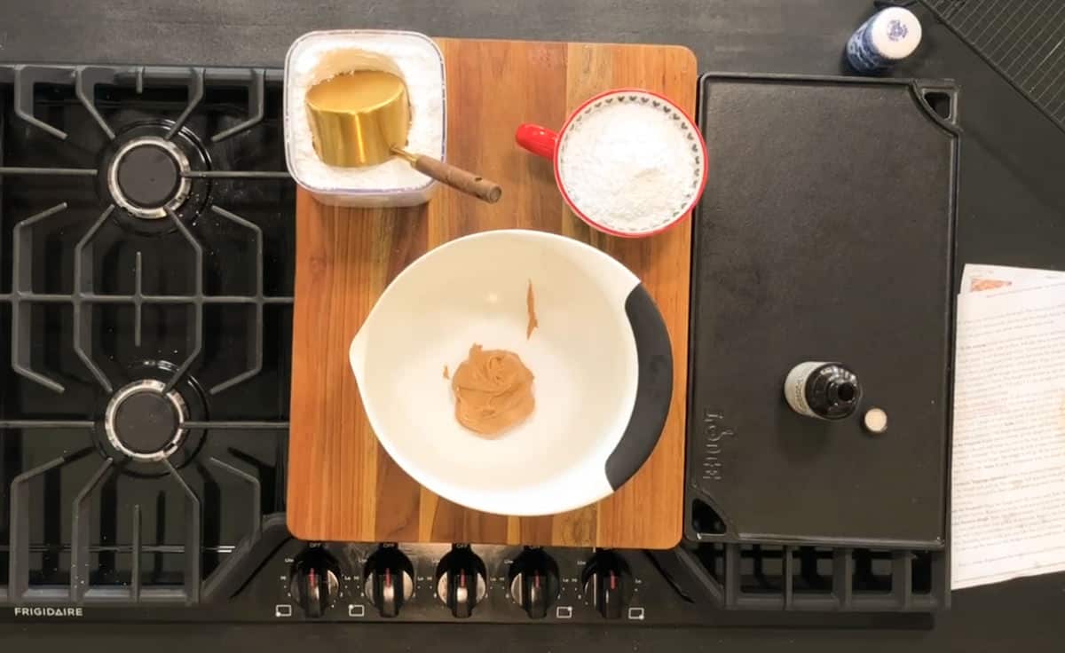 A kitchen countertop setup with a gas stove. In the center, a wooden board holds baking ingredients: a bowl with peanut butter, a cup with flour, and a container with a measuring cup inside. Nearby is a bottle of vanilla extract and a piece of paper.