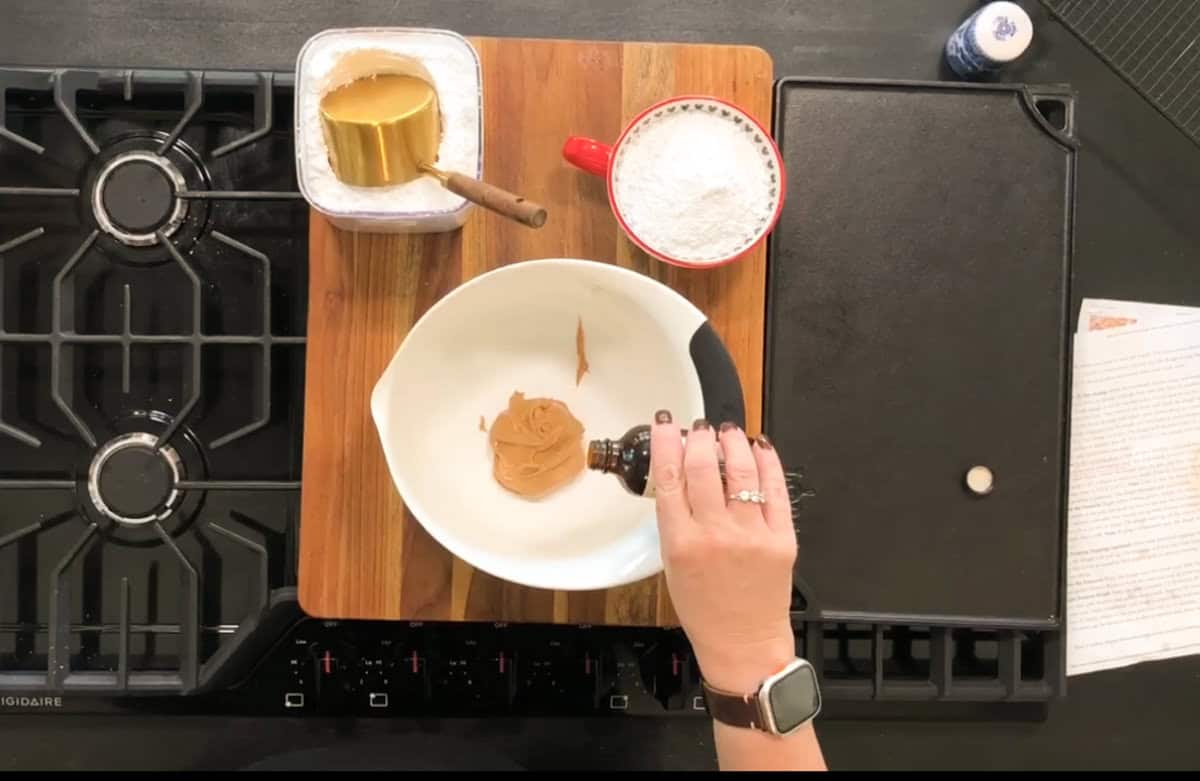 A person is pouring vanilla extract into a bowl containing peanut butter. Nearby are a measuring cup with flour and a container with a measuring cup filled with brown sugar, all on a wooden cutting board next to a stove.