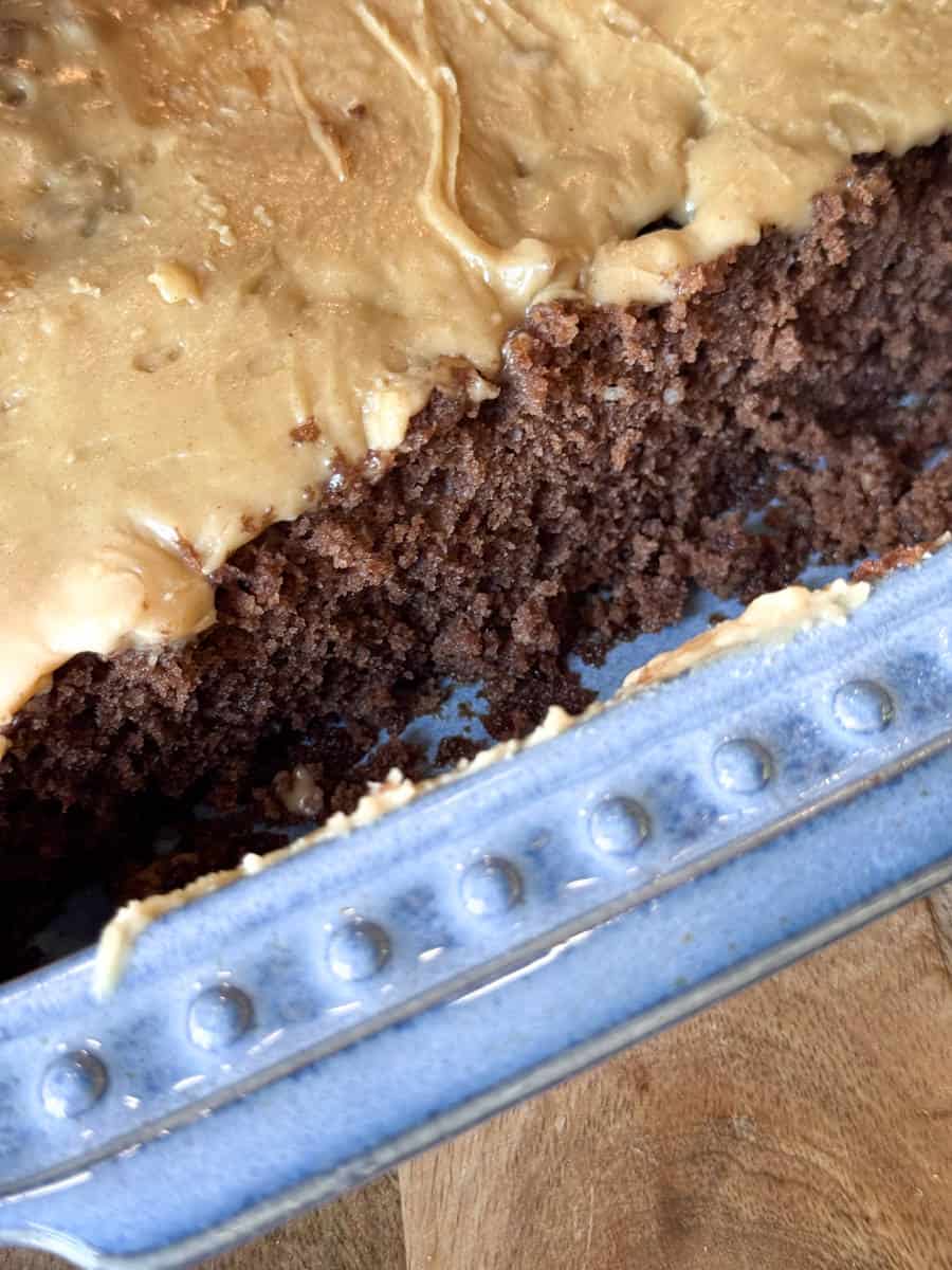 A close-up of a chocolate cake in a blue ceramic dish. The cake has been partially sliced, revealing its moist texture. It is topped with a thick layer of creamy brown frosting. The dish rests on a wooden surface.
