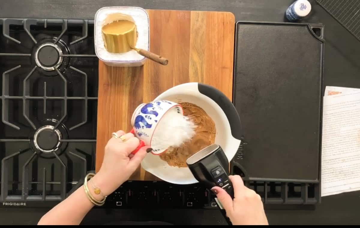 A person is using an electric hand mixer to blend ingredients in a white bowl placed on a wooden cutting board. They are adding sugar from a patterned ceramic cup into the bowl. The setup is on a stove, with a jar and paper nearby.