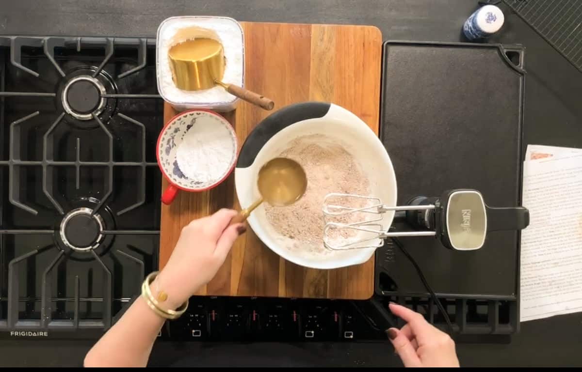 A person is preparing a mixture in a white bowl on a wooden cutting board. An electric mixer, a gold-colored measuring cup, and a red measuring cup with flour are nearby. The scene is set on a black stovetop surface.